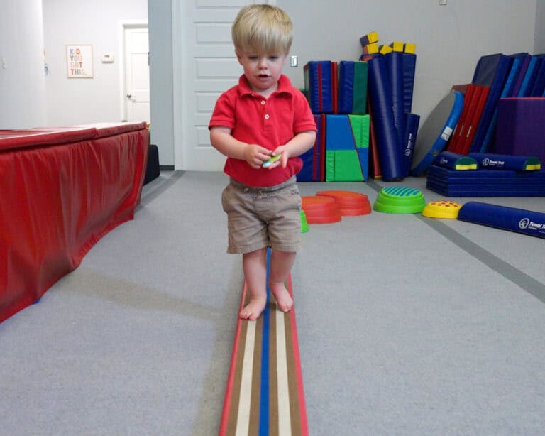 A photo of a boy at an OT clinic walking on the balance beam while holding an object for a balance challenge.
