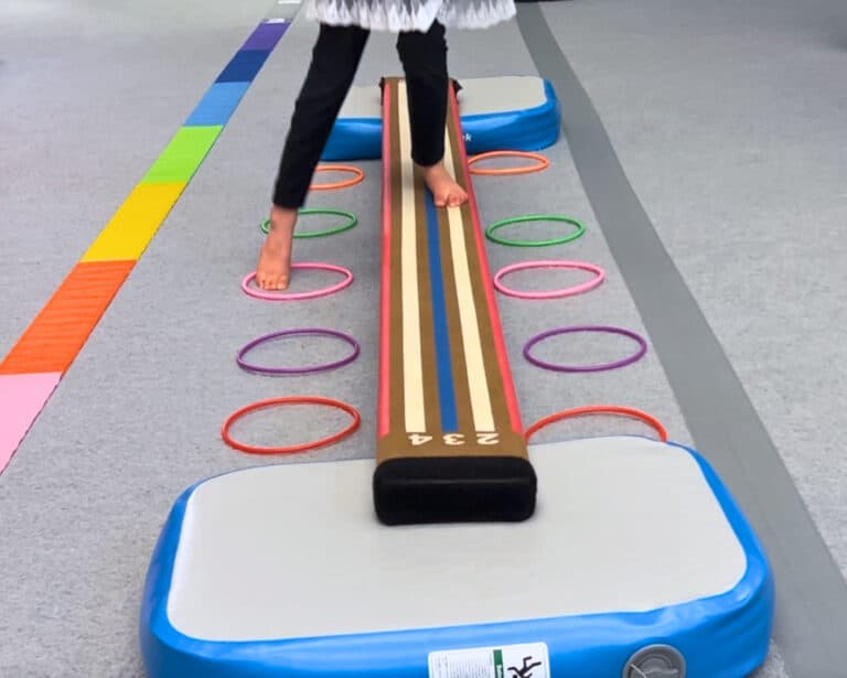 A photo in a pediatric OT clinic showing a client walking with one foot on the Laser Balance Beam and the other touching rings alongside the beam.