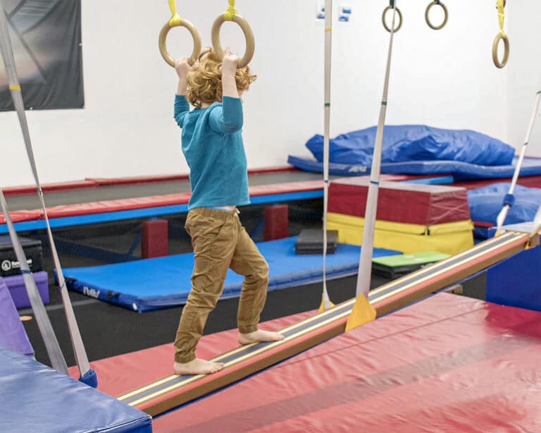 A photo of a boy walking up a Laser Beam Hammock suspended on an incline. He is reaching for Movement Rings above him.