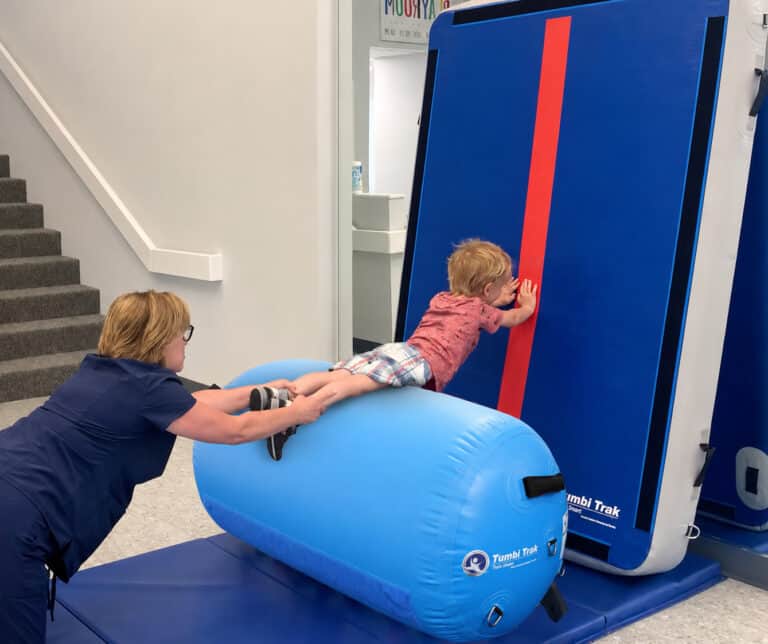 A photo of a boy in an OT clinic supported by an Air Barrel and pressing against a Power Launch that's stood up against a wall.