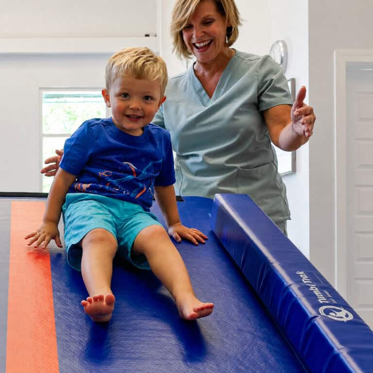 A photo of a boy and his pediatric OT smiling at the camera while he is sliding down a Power Launch.