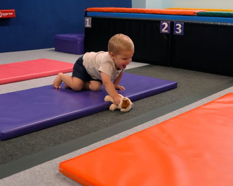 A photo of a boy crawling across a Build-a-Mat, holding a stuffed animal.