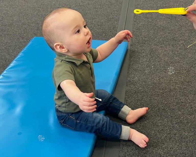A toddler sits on the edge of a Build-a-Mat, watching an OT with a bubble wand.
